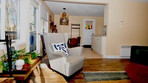 Una sala de estar con un sillón estampado, un sofá rojo y una mesa de centro de madera con plantas. Se observa una cocina al fondo.