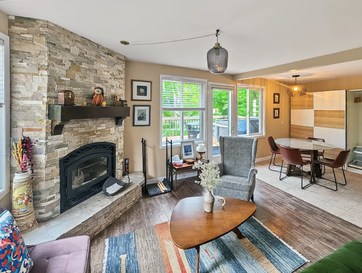 A living room with a stone fireplace, a dining area with a table and chairs, and large windows with a view of greenery.