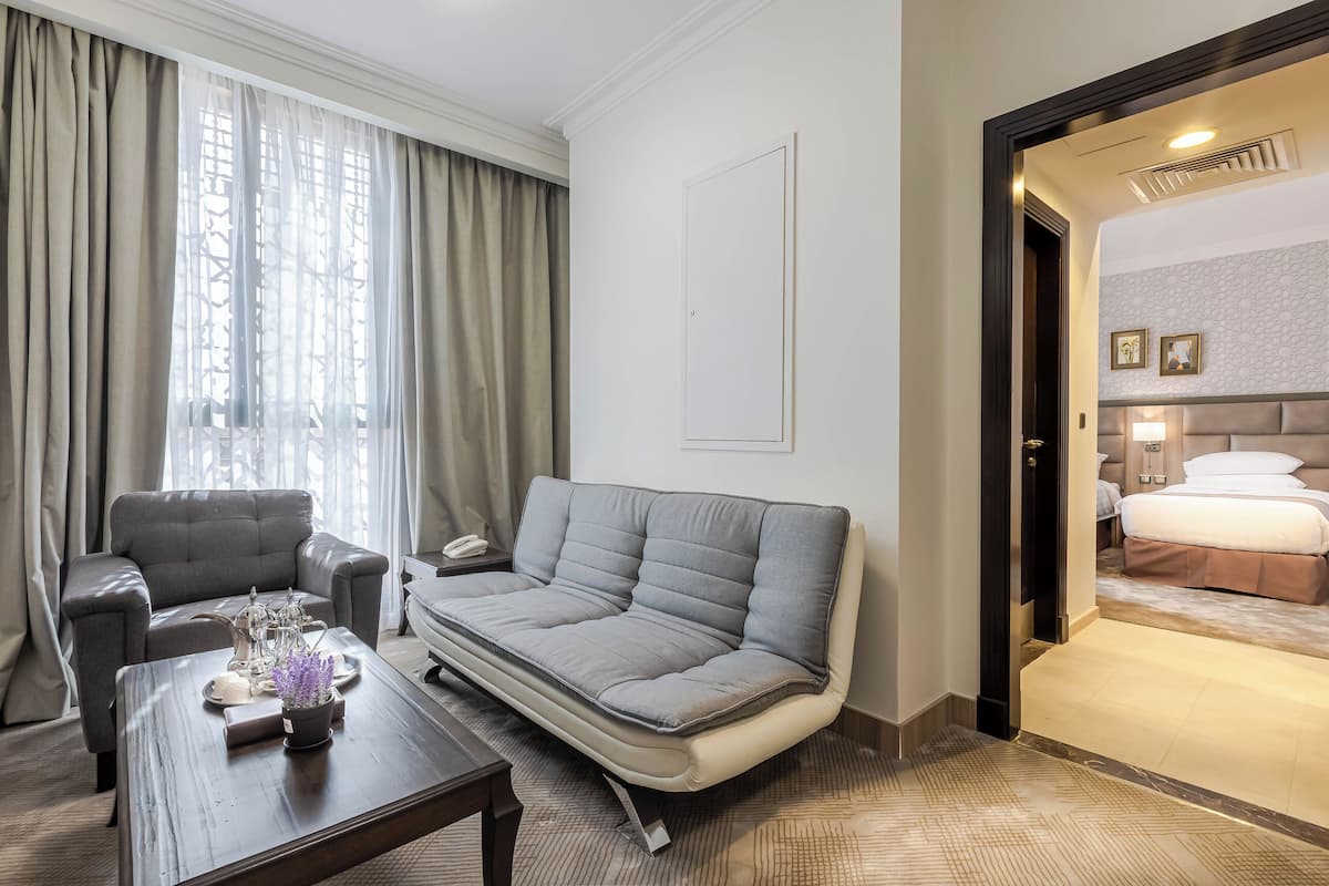 Habitación de hotel con un sofá gris, una mesita con un florero, una cama con cabecero y vistas a través de un vano.