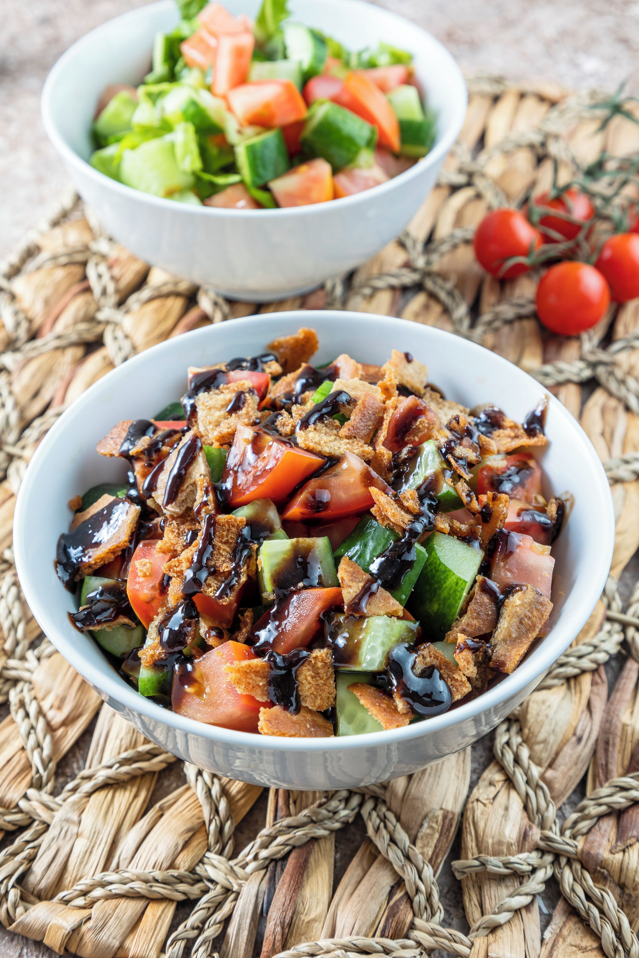 Eine Salatschüssel mit Tomaten, Gurken und einem dunklen Dressing, auf einer gewebten Matte platziert.
