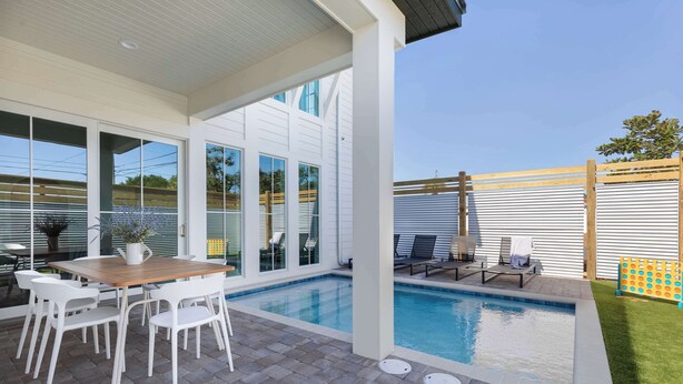 A modern outdoor pool area with a wooden table and chairs, a covered patio, and a fenced yard.