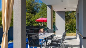 Un patio exterior con una mesa y sillas, una sombrilla roja y una piscina al fondo.
