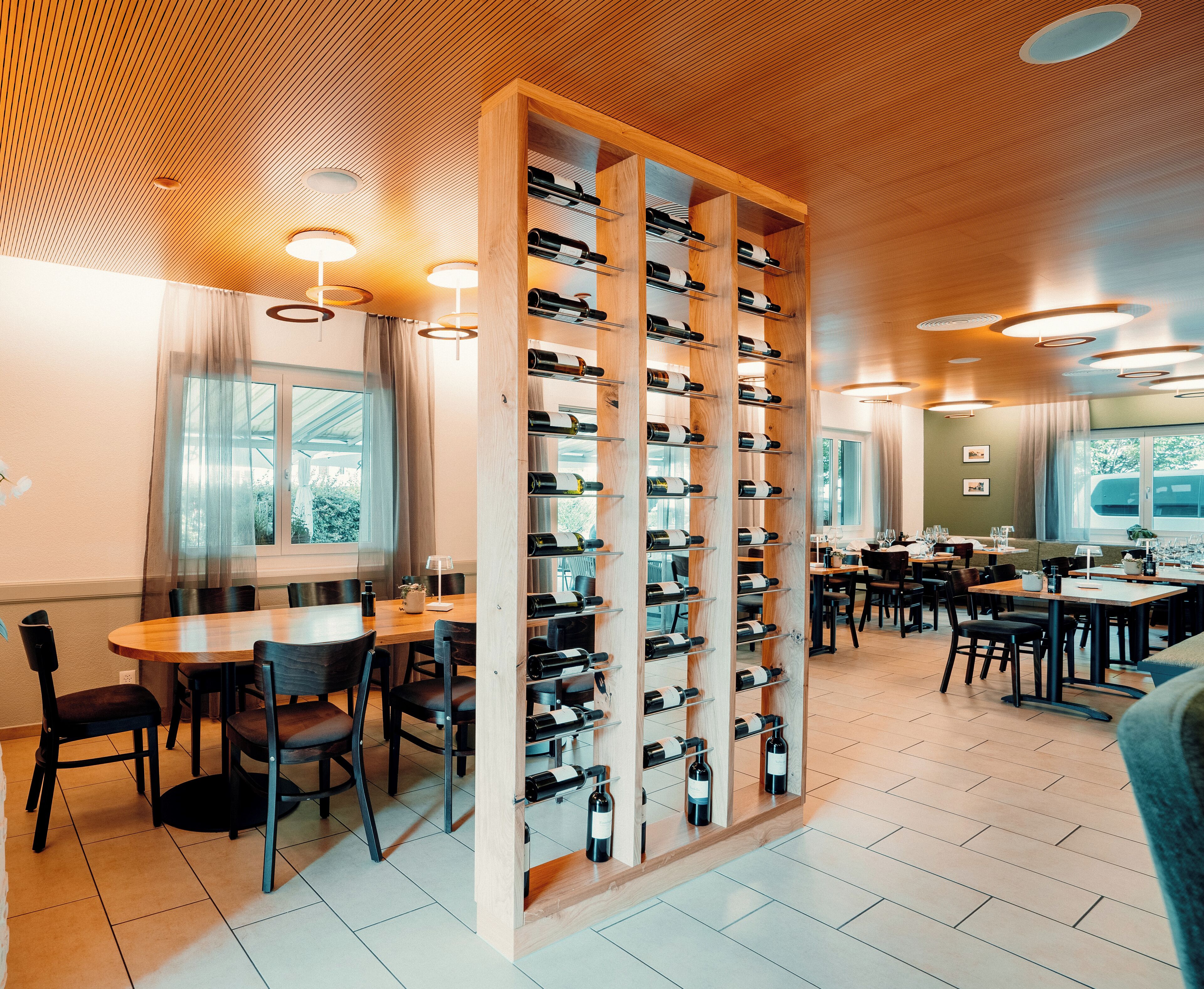 Interior de un restaurante con estanterías de madera para vino, mesas, sillas e iluminación colgante.