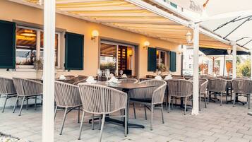 Een terras buiten met meerdere tafels en stoelen, een overdekte zithoek en uitzicht op de straat.