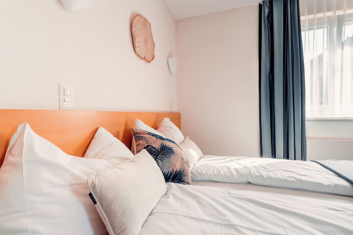 Un lit bien fait, avec du linge de lit blanc et une tête de lit en bois, un luminaire fixé au mur et une fenêtre avec des rideaux.