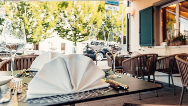 Een terras met eettafels voor twee personen, compleet met wijnglazen en bestek. In de achtergrond is een 'HOTEL' bord te zien.