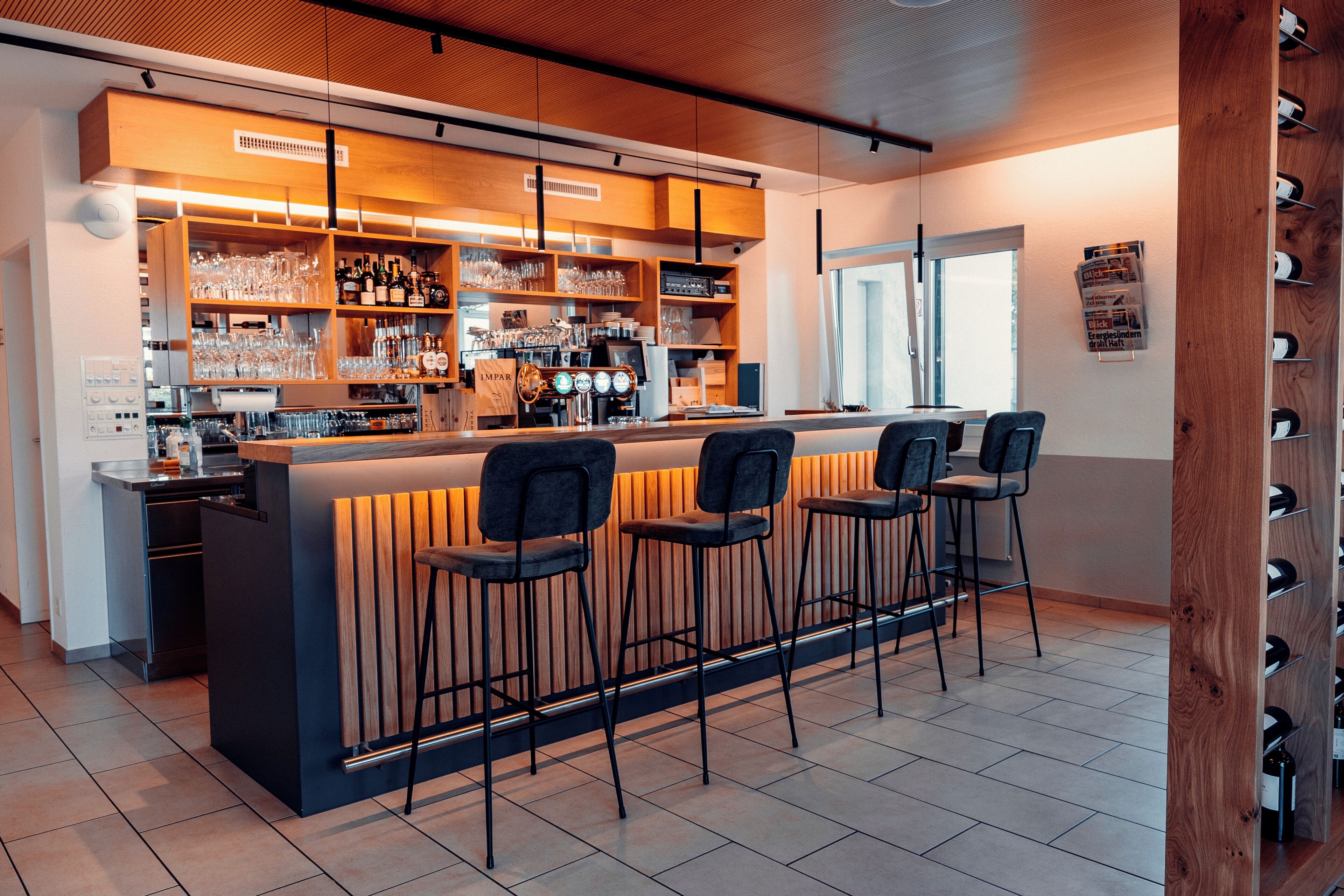 A modern bar area with a wooden bar counter, multiple stools, and a wine rack.