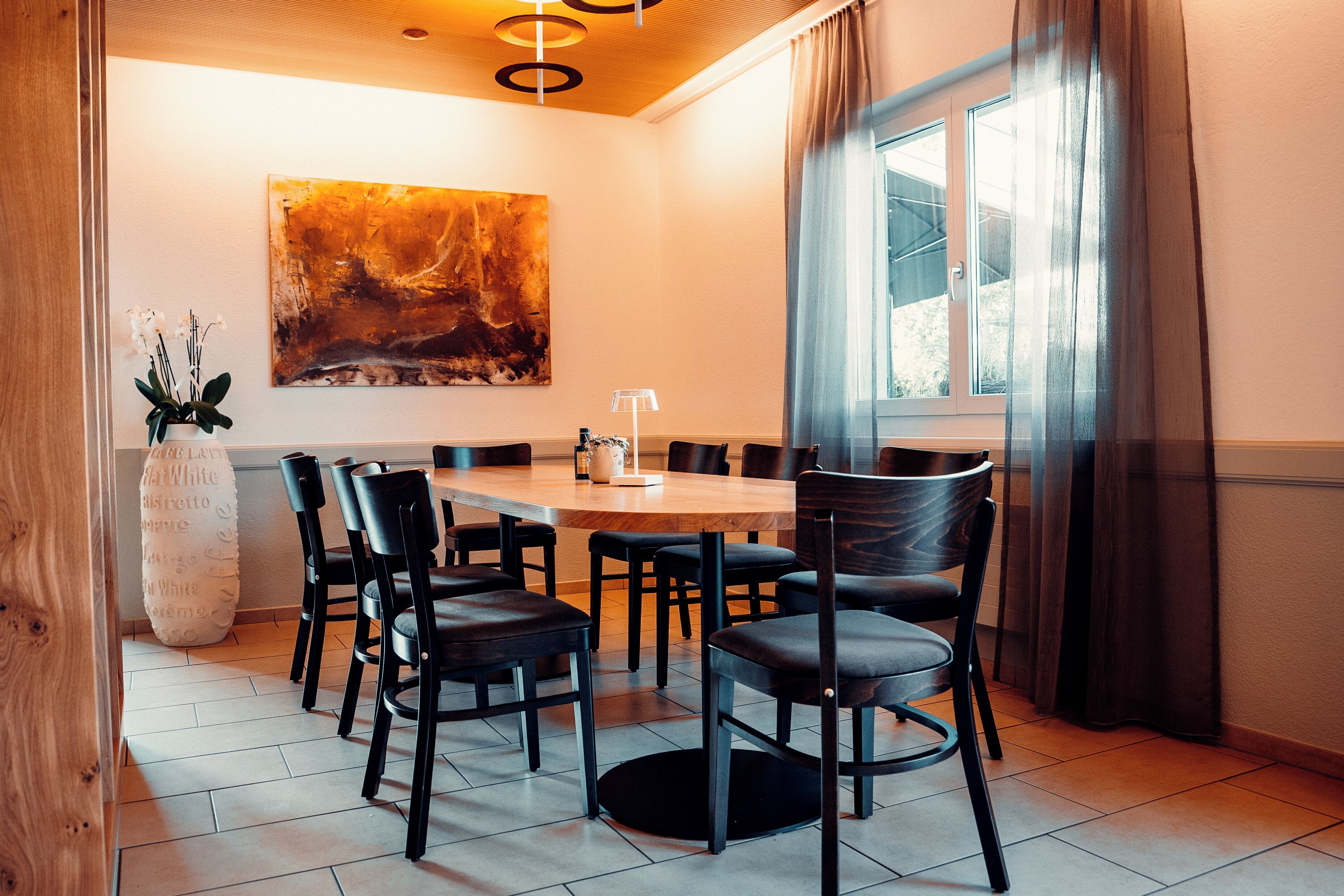 A dining area with a wooden table and black chairs, a large abstract painting on the wall, and a window with curtains.