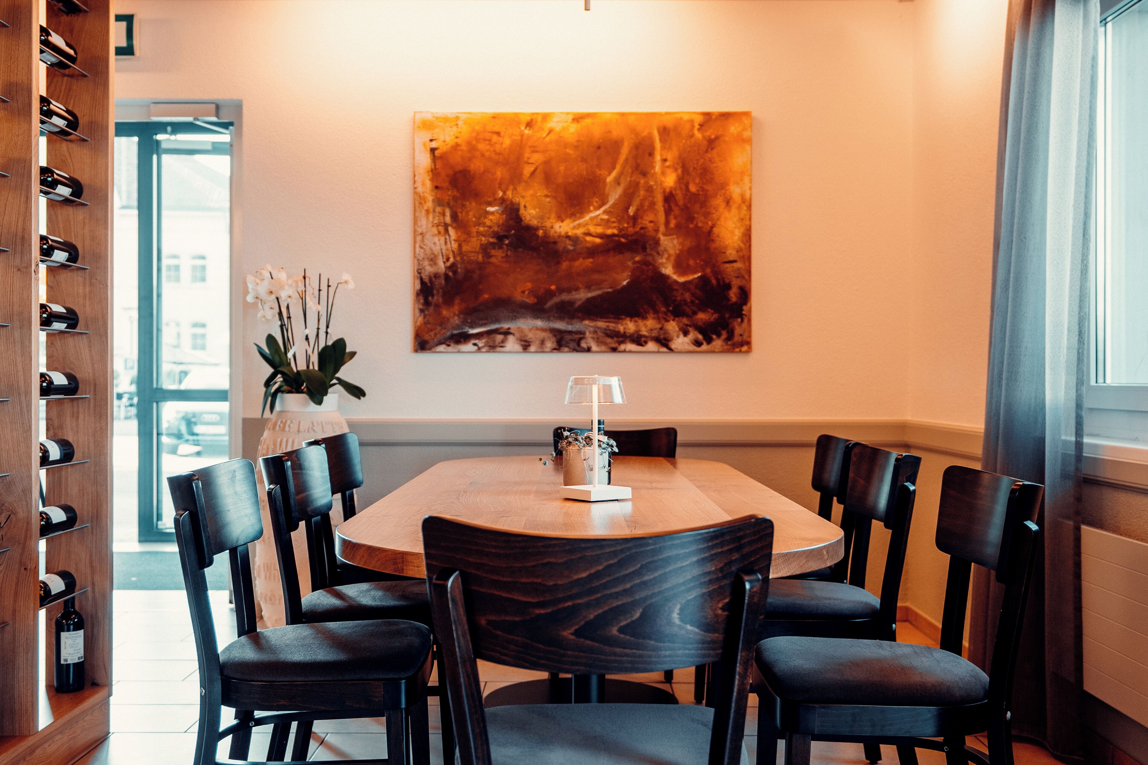 A dining area with a round table, six chairs, a wine rack, and a large abstract painting on the wall.