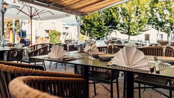 Un espacio de comedor al aire libre con mesas dispuestas para dos, con sillas de madera oscura y un toldo en la parte superior.
