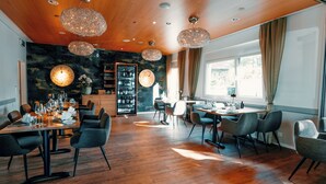 Een moderne eetruimte met houten vloeren, ronde tafels en moderne stoelen. In de achtergrond hangen kristallen kroonluchters en is een grote wijnkast zichtbaar.