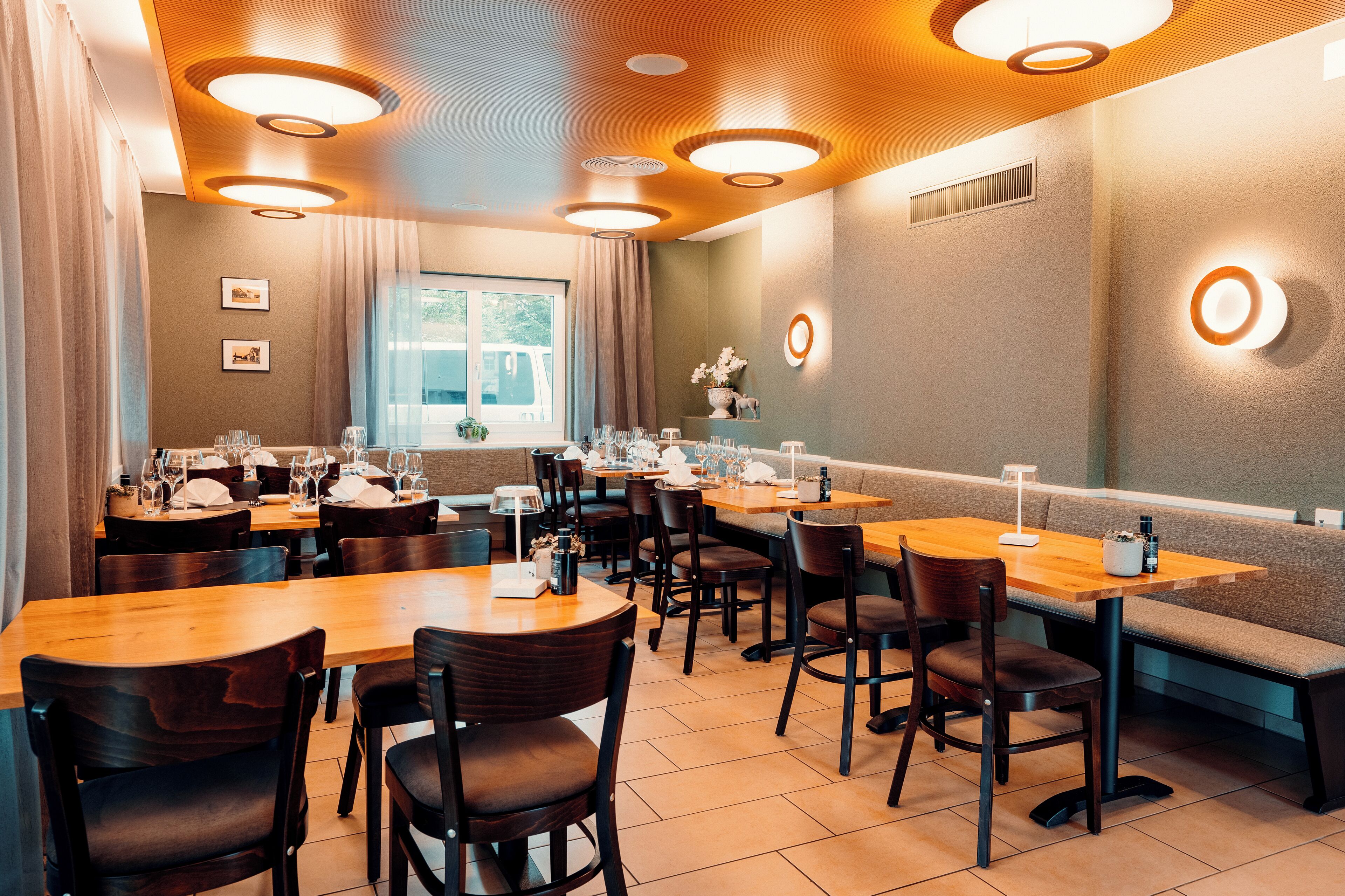 Un restaurante con mesas y sillas de madera, un amplio ventanal e iluminación en las paredes.