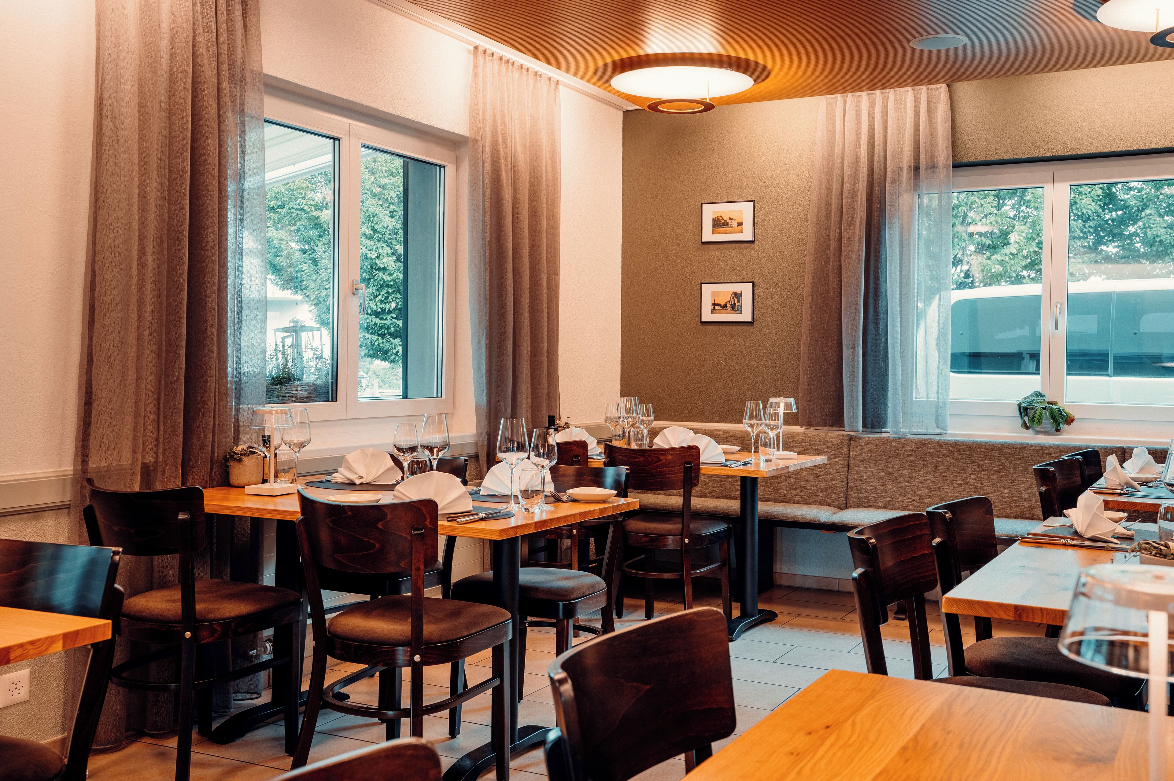 A restaurant with wooden tables and chairs, a bench seating area, and large windows with curtains.