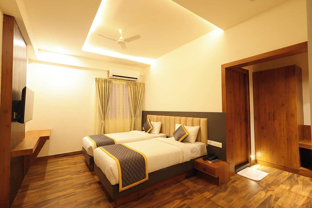 A hotel room with two beds, wooden flooring, a ceiling fan, an air conditioning unit, a TV, and a wardrobe.