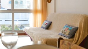 Una habitación con un sofá color beige, dos almohadas, una mesa con un vaso y una ventana con cortinas.