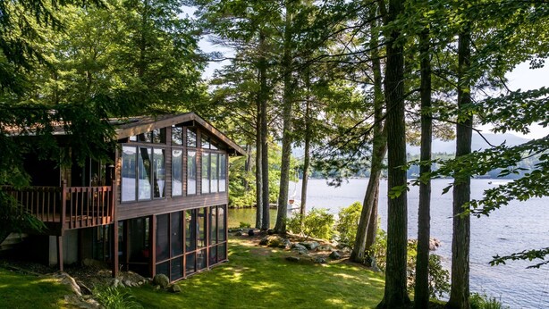 Una cabaña con terraza y vistas a un lago, rodeada de árboles.