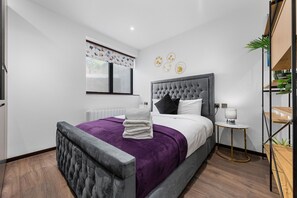 A modern bedroom with a tufted headboard, a large window with blinds, a bedside table with a lamp, and a small shelf with plants. - Bright Studio Retreat in Golders Green (London)