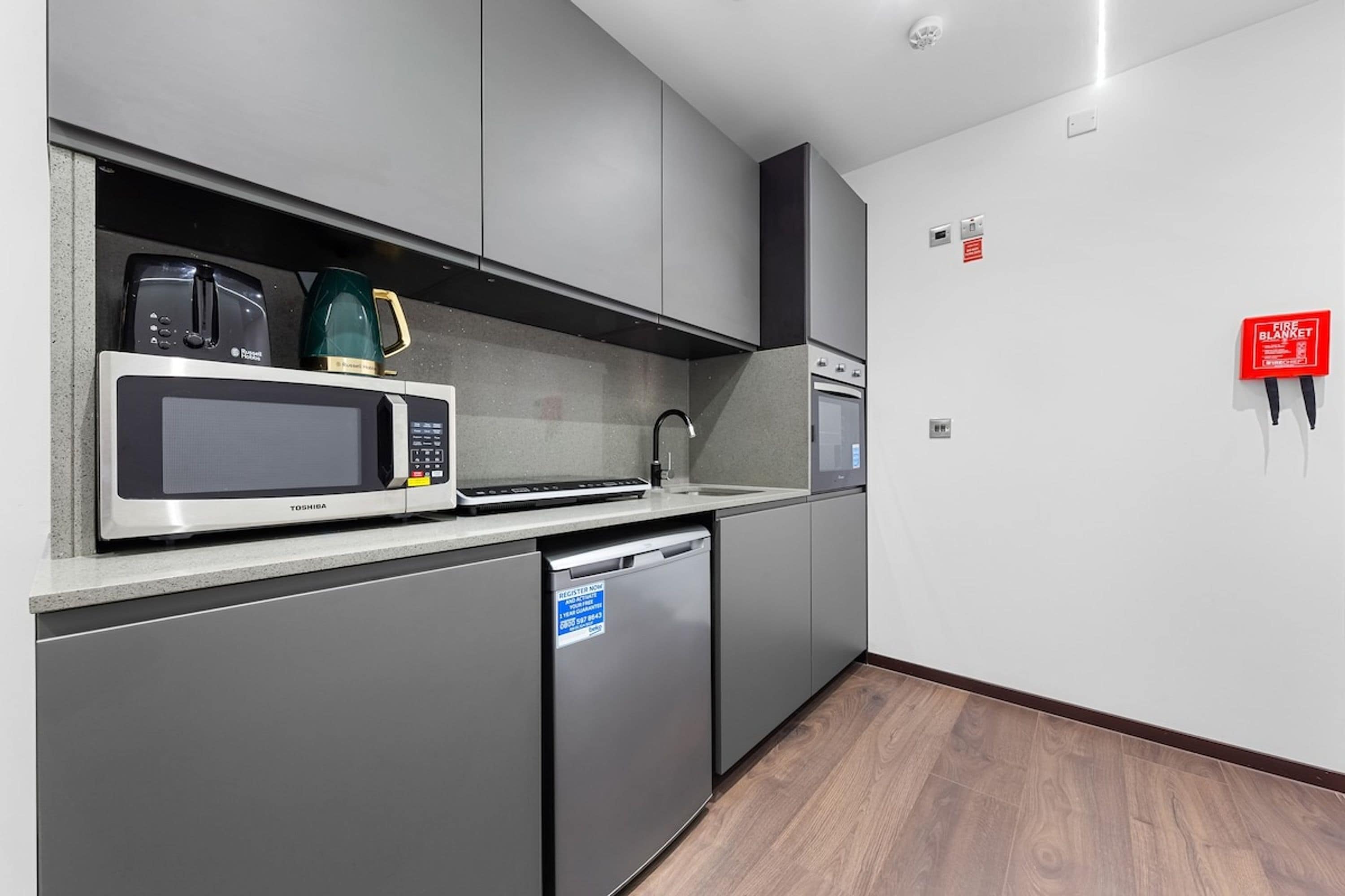 A modern kitchen with a microwave, toaster, and coffee maker on the countertop, a dishwasher, and a red fire alarm on the wall.