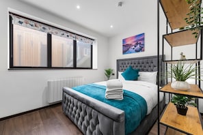 A modern bedroom with a large bed, a grey upholstered headboard, a teal bedspread, and a window with blinds.