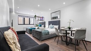 A modern hotel room with a bed, a sofa, a dining table with chairs, a mirror, and a window with blinds. - Cosy Golders Green Studio With Modern Amenities (London)