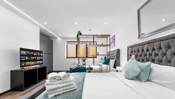 A modern hotel room with a large bed, a flat-screen TV, a seating area with a table and chairs, and a small balcony. - Cosy Golders Green Studio With Modern Amenities (London)