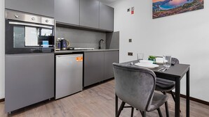 A modern kitchen with grey cabinets, a built-in oven, and a small refrigerator. There is a dining table with chairs and a painting of a sunset on the wall. - Chic Studio in Golders Green Near Tube Station (London)