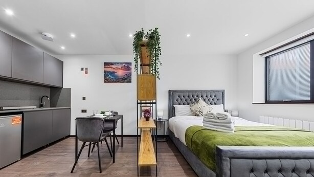 A modern bedroom with a large bed, a bedside table, a desk, and a chair. There is a window with blinds and a painting on the wall. - Chic Studio in Golders Green Near Tube Station (London)