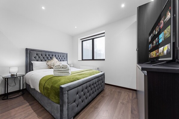 Une chambre moderne avec une tête de lit capitonnée, un plaid vert, un lit soigneusement fait, une table de chevet avec une lampe, une grande fenêtre avec des stores et un téléviseur fixé au mur.