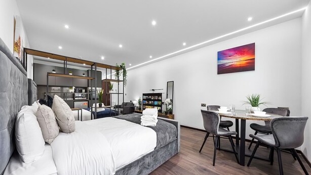 A modern hotel room with a bed, a dining table with chairs, a kitchen area, and a large window. - Stylish Studio in Golders Green Central Location (London)