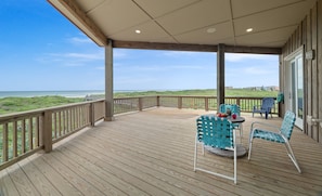 Une terrasse en bois offrant une vue sur la plage, des meubles d’extérieur et une porte vitrée donnant sur l’intérieur.