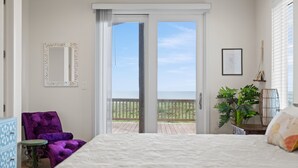 A bedroom with a large bed, a purple chair, a small table, a potted plant, and a balcony with a view.