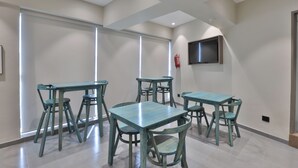 Ένας σύγχρονος χώρος τραπεζαρίας με πολλά τραπέζια και καρέκλες, μια τηλεόραση επίπεδης οθόνης τοποθετημένη στον τοίχο και ένα μεγάλο παράθυρο που παρέχει φυσικό φως.