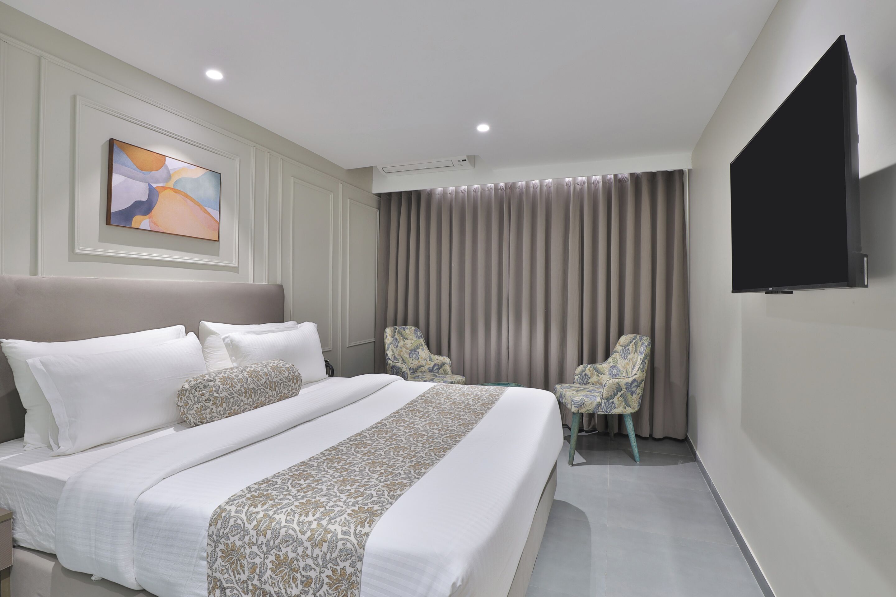 A modern hotel room with a large bed, two armchairs, a flat-screen TV, and a painting on the wall.