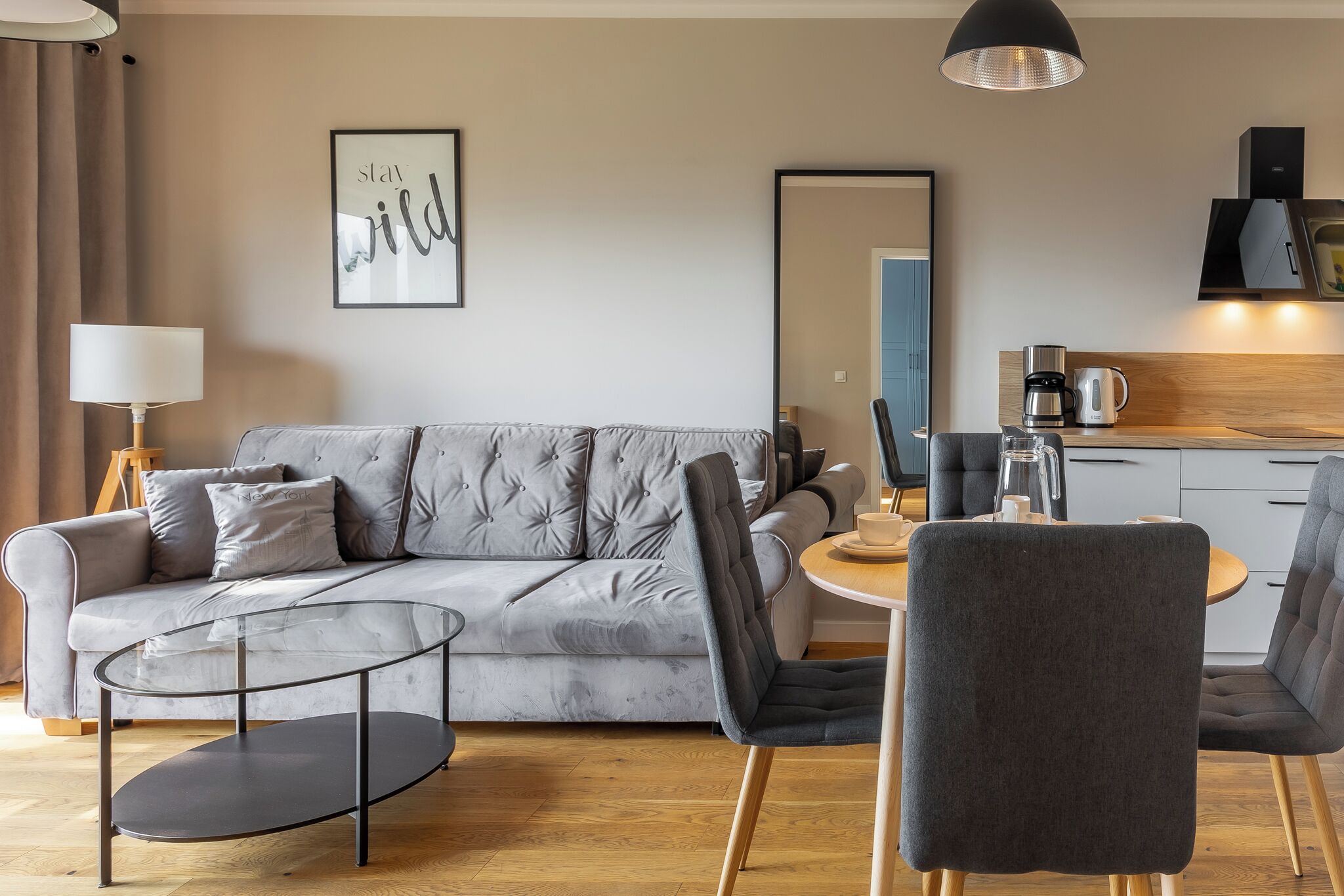 Ein modernes Wohnzimmer mit grauem Sofa, einem Glastisch und einem runden Esstisch mit Stühlen. Ein großer Spiegel hängt an der Wand, und im Hintergrund ist eine Küchenzeile erkennbar.