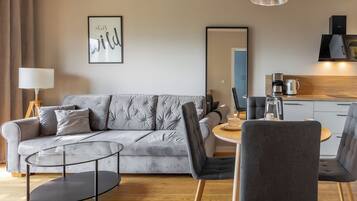 Ein modernes Wohnzimmer mit grauem Sofa, einem Glastisch und einem runden Esstisch mit Stühlen. Ein großer Spiegel hängt an der Wand, und im Hintergrund ist eine Küchenzeile erkennbar.
