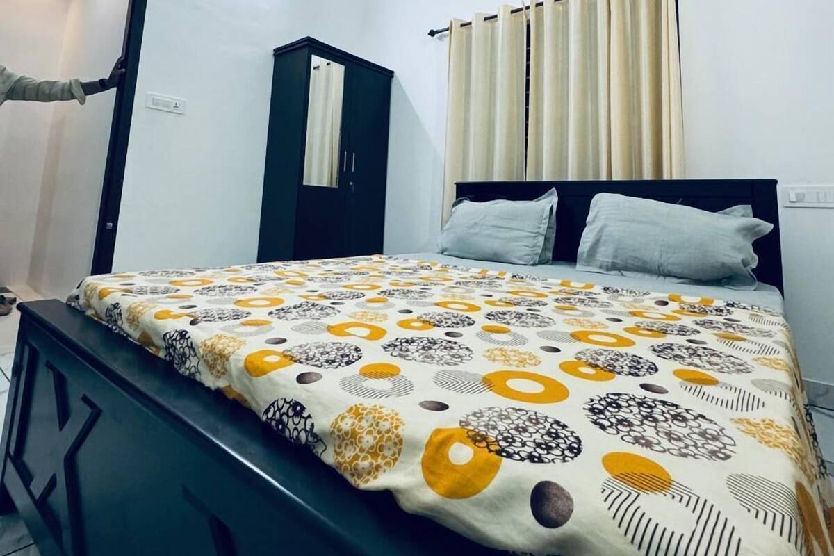 A bedroom with a bed, a wardrobe, a mirror, a window with curtains, and an air conditioning unit.