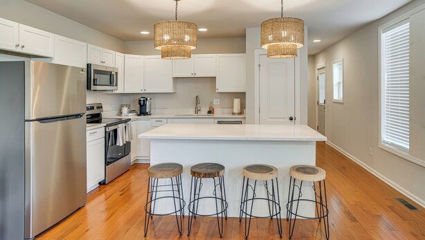 A modern kitchen with white cabinets, stainless steel appliances, a large island with stools, and wicker pendant lights. - Walk to Five Points: East Nashville Home w/ Yard! (Nashville)