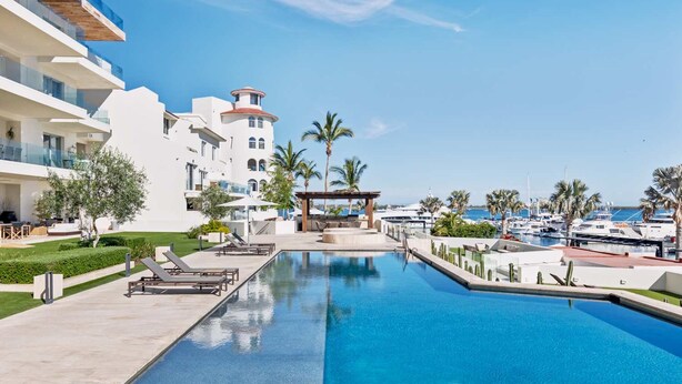 Ein Poolbereich eines Resorts mit freiem Blick auf das Meer, Palmen und einem Yachthafen im Hintergrund.
