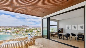Ein modernes Zimmer mit Holzboden, großen Fenstern, einem Balkon mit Blick auf Strand und Stadt sowie einem Essbereich mit Stühlen und Tisch.