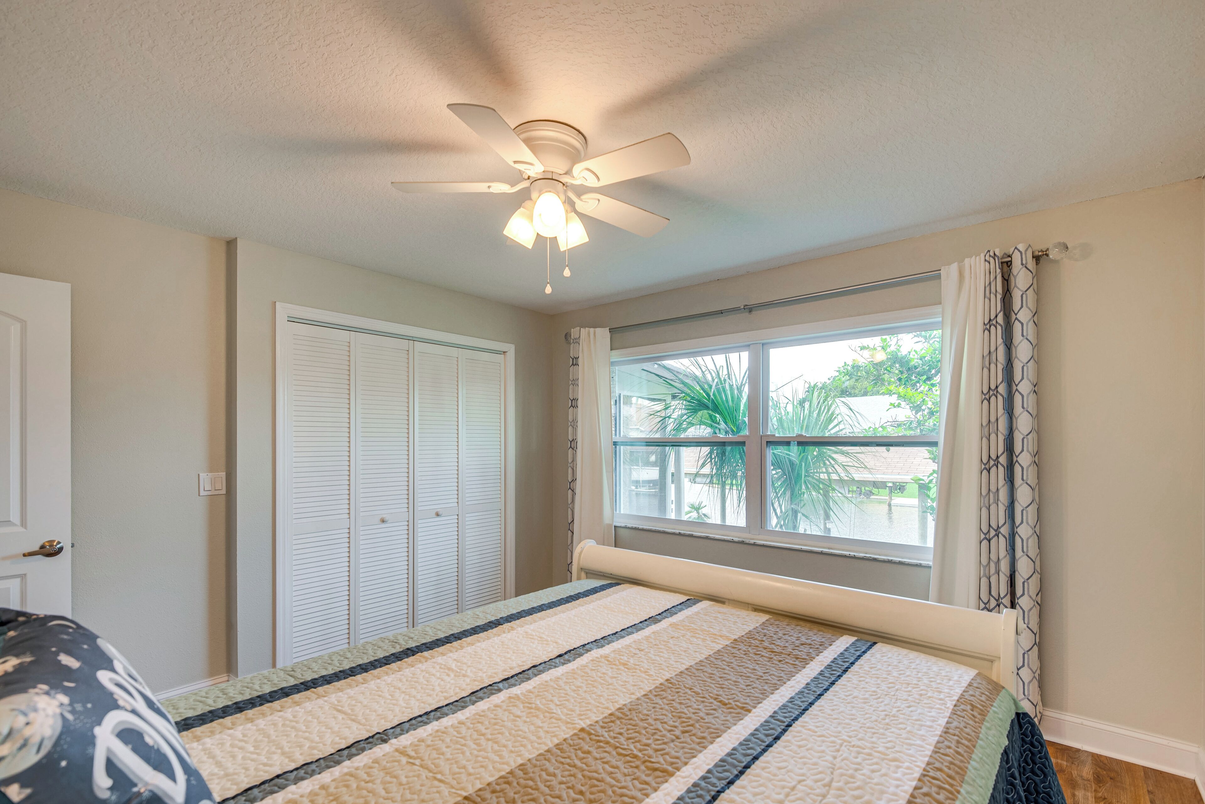 Un dormitorio con una cama, un ventilador de techo, una ventana con cortinas y una puerta.