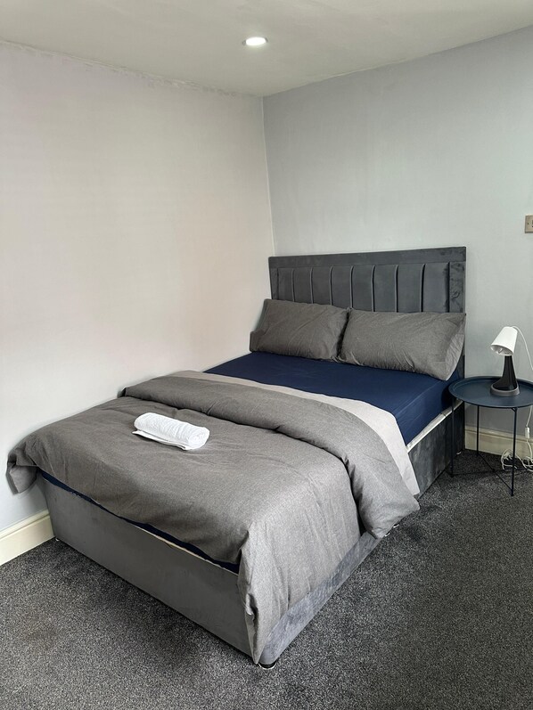 A neatly made bed with grey bedding, a dark headboard, and a small bedside table with a lamp. - Cozy Manchester House - Stay With Host Onsite (Stockport)