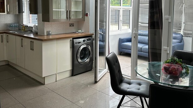 Une cuisine moderne équipée d’un lave-linge intégré, une table à manger en verre avec des chaises, et une vue sur un espace extérieur aménagé avec des sièges.