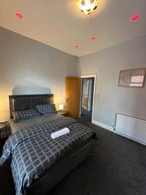 A bedroom with a large bed, a nightstand, a lamp, and a door leading to another room. - Cozy Manchester House - Stay With Host Onsite (Stockport)