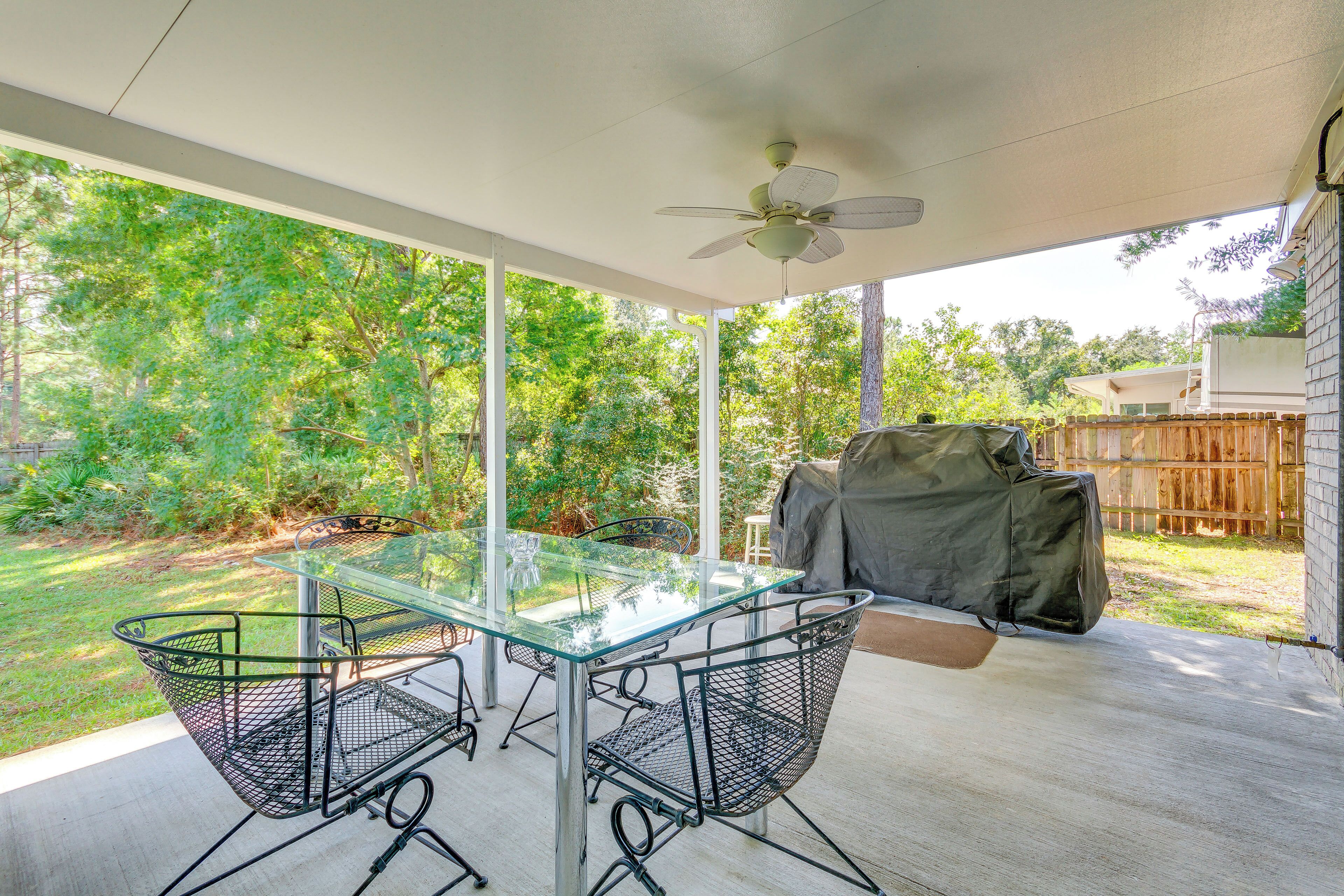 A covered patio with a glass table and metal chairs, a ceiling fan, and a view of a backyard with trees and a wooden fence.
