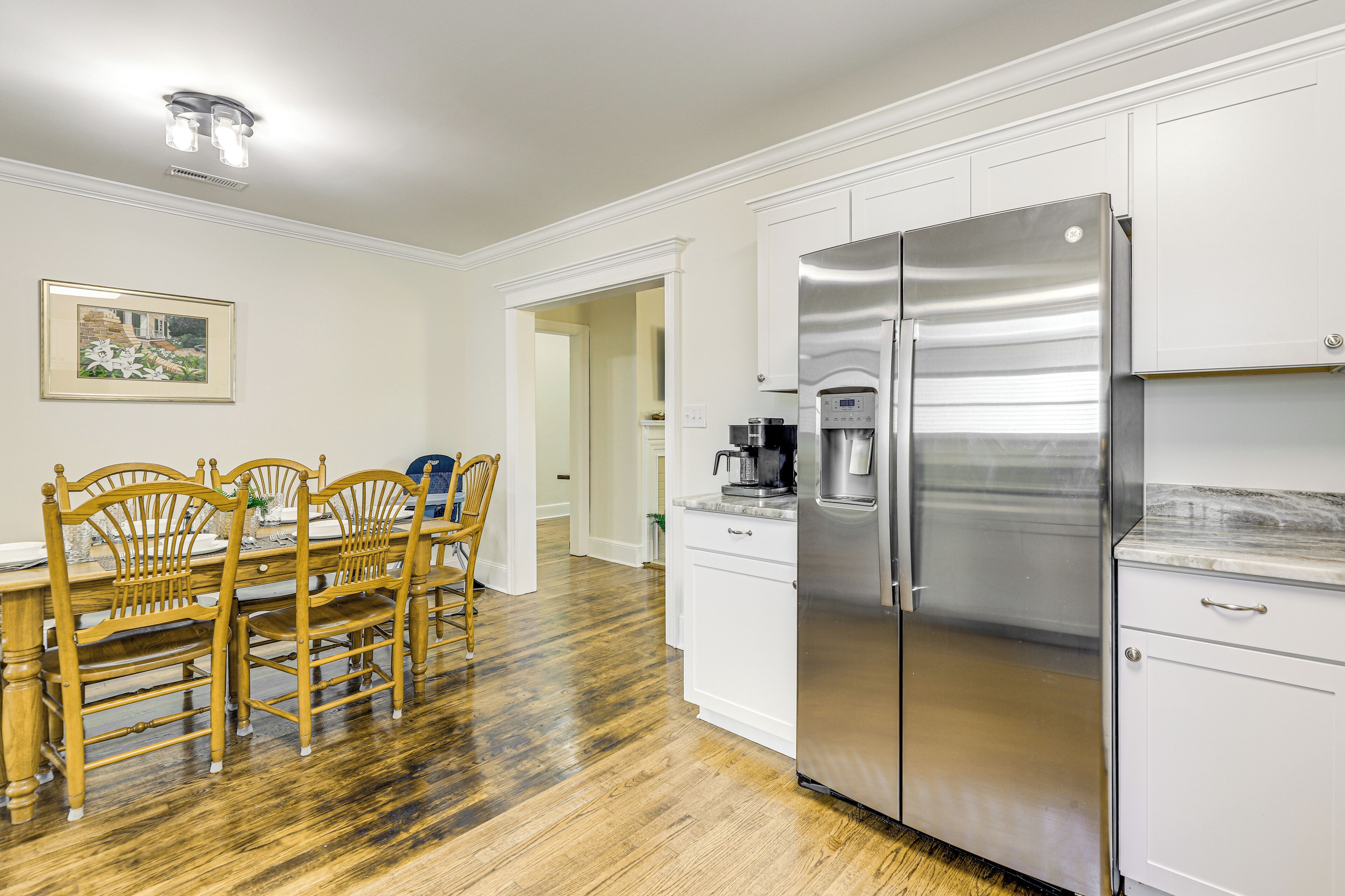 A kitchen with white cabinets, a stainless steel refrigerator, a wooden dining table with chairs, and a framed picture on the wall.