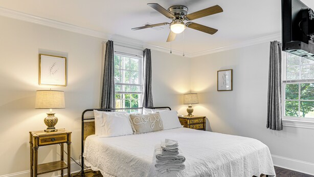 Habitación de hotel con cama, dos mesitas de noche con lámparas, ventilador de techo y ventana con cortinas.