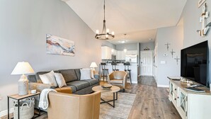 A modern living room with a sofa, coffee table, and a TV. There is a kitchen in the background with white cabinets and a dining area.
