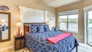 A bedroom with a large bed, a nightstand with a lamp, a dresser, and a view of a lake through a window. - Lakefront Camdenton Condo Near Ha Ha Tonka (Camdenton)
