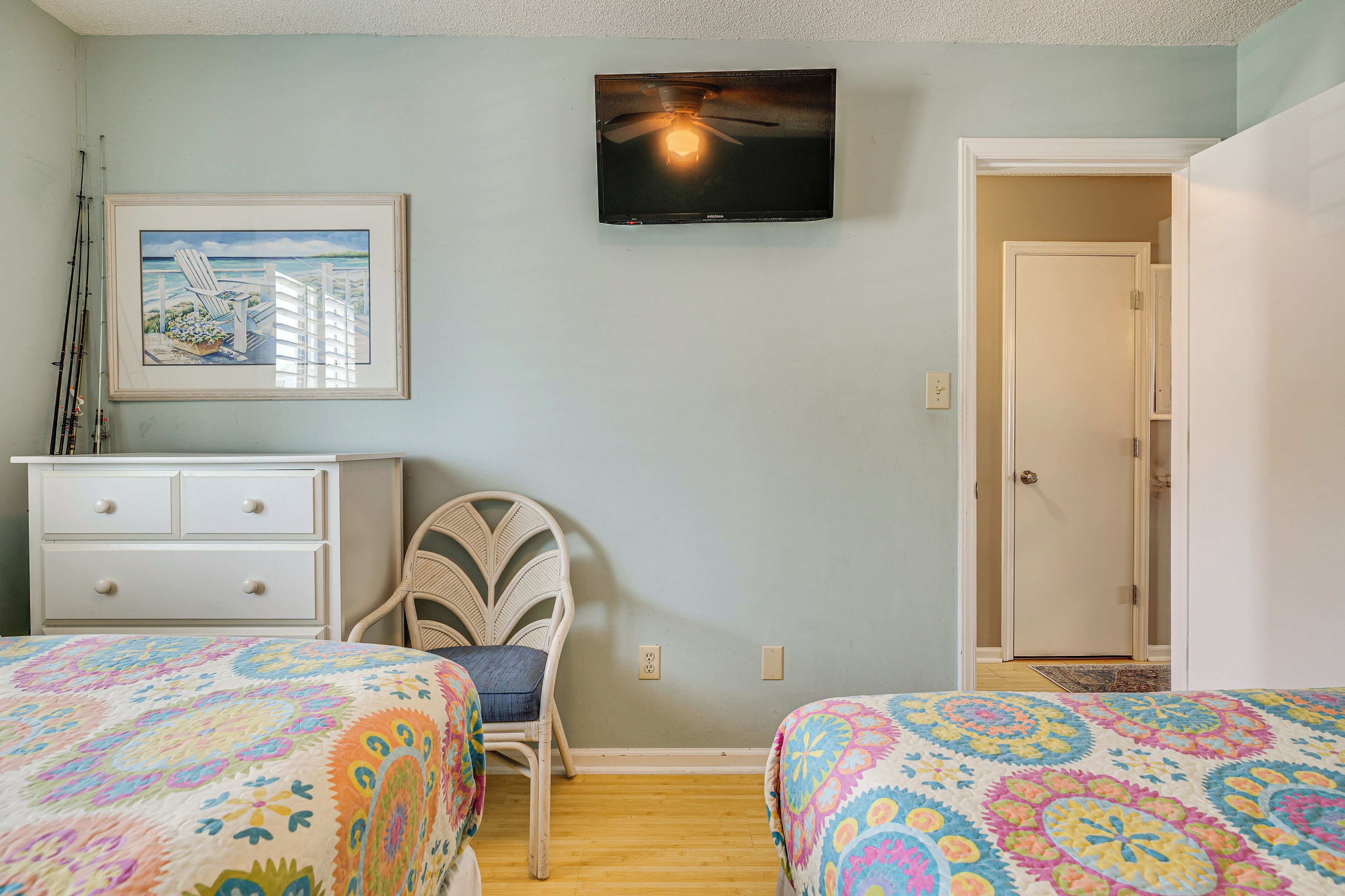 Une chambre avec deux lits, un ventilateur de plafond, une télévision, un tableau encadré, une commode et une chaise.