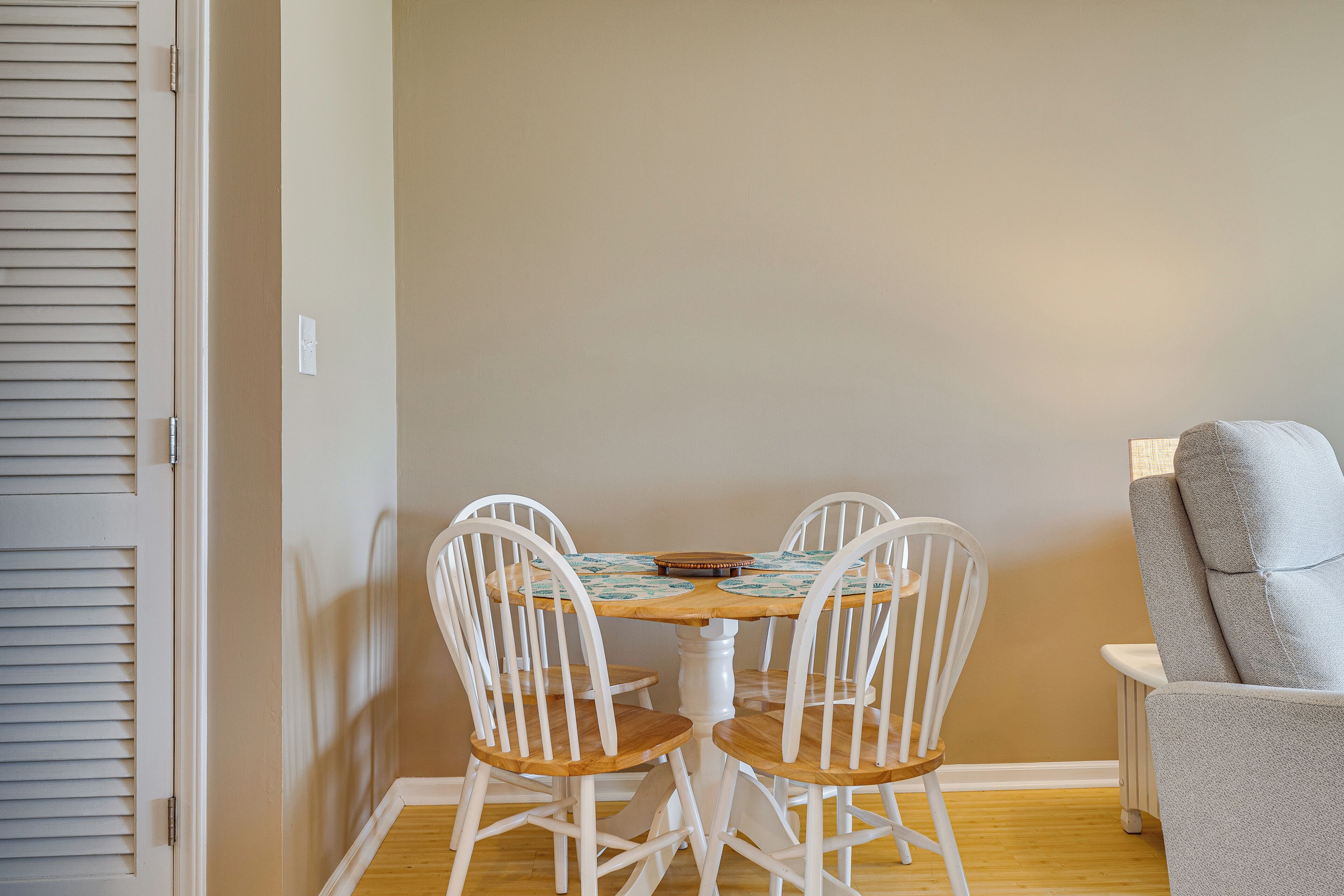 A dining area with a table and four white chairs, a grey armchair, and a closed door with blinds.